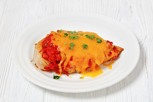 baked chicken breasts topped with finely chopped tomatoes, cheddar cheese and parsley on white plate on white wooden table
