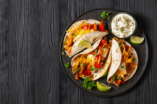 baked tex-mex chicken fajitas with mixed sweet pepper, onion, sour cream, shredded cheese and white corn tortillas on black plate on black wooden table, horizontal view,  flat lay, copy space