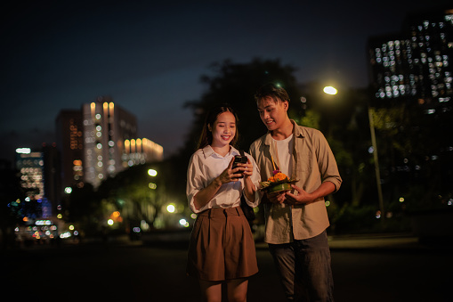 Smiling couple walks together at the Loy Krathong festival night, and looks at the phone.