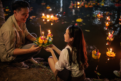 A smiling couple holds a krathong by the river during the Loy Krathong festival night.