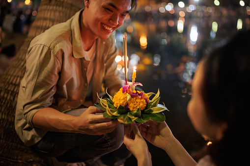 A smiling couple holds a krathong by the river during the Loy Krathong festival night.