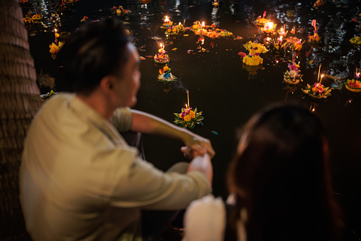 A couple is watching the floating krathongs while sitting by the river on the night of the Loy Krathong.