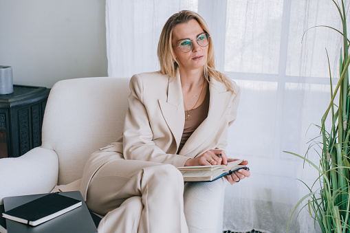 Professional woman in a beige suit thoughtfully reading a book in a well-lit room, with sheer curtains and a plant adding to the serene ambiance
