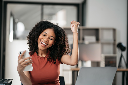 Overjoyed charming excited black woman business woman worker using smartphone and laptop working in office, feeling happy.