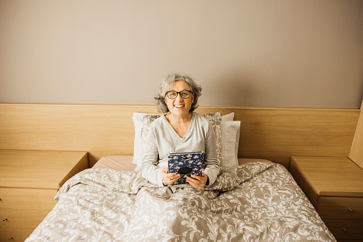 Portrait of a smiling senior woman sitting on bed with pajama and looking at her digital tablet.