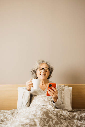 Senior woman sitting on bed in the morning with pajama and drinking coffe with a white mug while checking her phone.