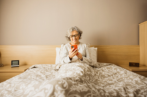 Senior woman sitting on bed with pajama in the morning and checking her messages on the phone.