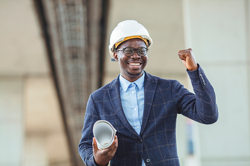 Portrait of an excited casual architect  standing with raised hands. Young architect man wearing contractor helmet holding project paper plan smiling and screaming for success. Celebration