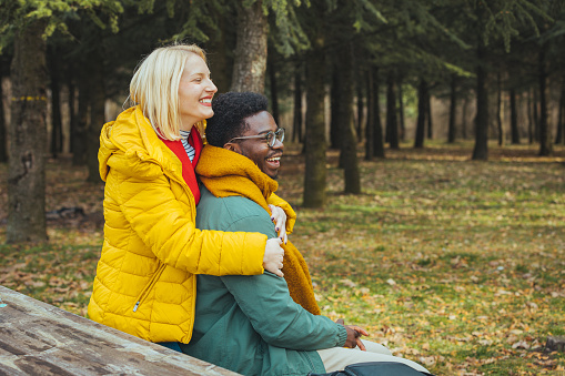 Young couple in love having fun and enjoying the beautiful nature. She hugs him from behind, sits on a bench in the woods and spends a beautiful time together.