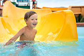 Cheerful blue-eyed kid on a water slide in the water park, a little boy merrily slides into the water along the slide in the amusement park, rest with children in the water park