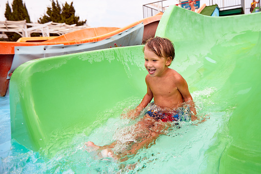Cheerful blue-eyed kid on a water slide in the water park, a little boy merrily slides into the water along the slide in the amusement park, rest with children in the water park.