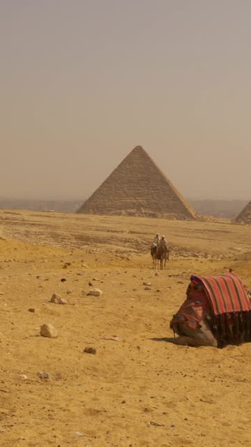 Cairo, Egypt; October 2020: Panning shot of two local men riding a camel next to the Ginza pyramids