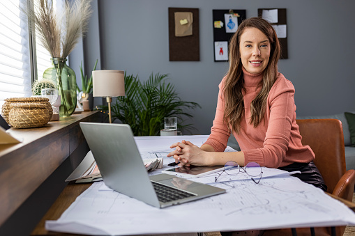 Smiling female entrepreneur. Female architect smiling to camera in open plan office. Female Architect Studying Plans In Office Smiling At Camera. Beautiful business woman sitting in the office, looking at camera and smiling