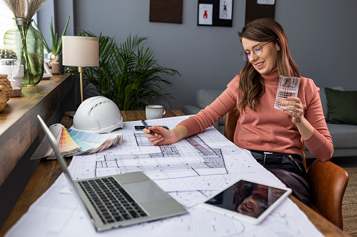 Female designer in office working on architects project and holding glass of water. Multitasking of young female entrepreneur at work. Young female entrepreneur drinking water at modern working place