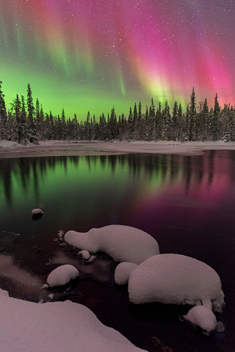 Winter night view with geomagnetic storm above the arctic sky showing northern lights with green and red colors reflecting in the water of a lake covered with snow and ice, Lapland, Finland, Scandinavia