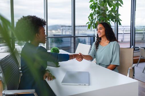 Two professional businesswomen, one Black and one Hispanic, engaging in a handshake over a meeting table in a well-lit office setting.