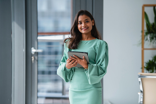 A professional Indian businesswoman holding a digital tablet, standing in a modern office environment.