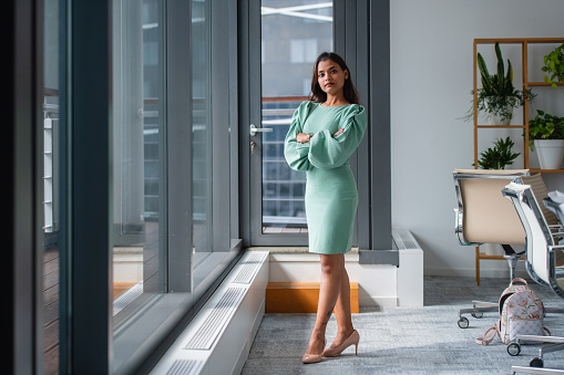 A professional young adult businesswoman stands with arms crossed in a well-lit modern office setting, exuding confidence.