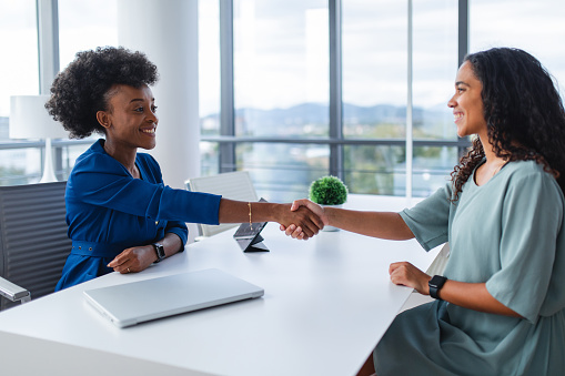 Two professional women shaking hands across a table in a corporate office environment, signifying agreement or introduction.