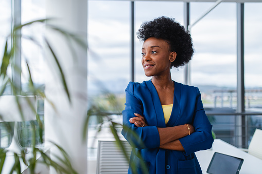 A professional black businesswoman in a blue blazer stands with arms crossed in a modern office, exuding confidence and leadership.
