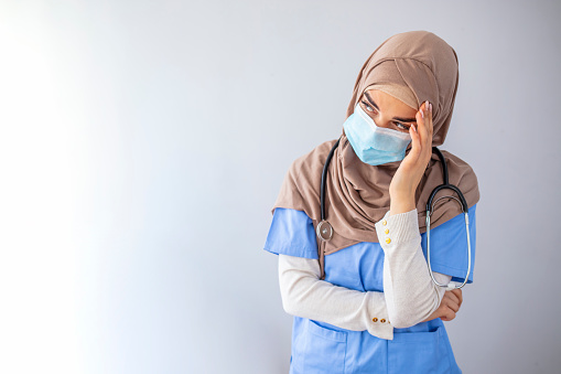 Stressed muslim woman doctor in protective face mask touching her sweating forehead, feeling exhausted after difficult working day, sitting by wall at clinic, copy space. Doctors and COVID-19 pandemic