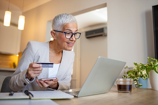 Mature woman using credit card making online payment at home. Successful old woman doing online shopping using laptop. Closeup of retired fashionable lady holding debit card for internet banking account.