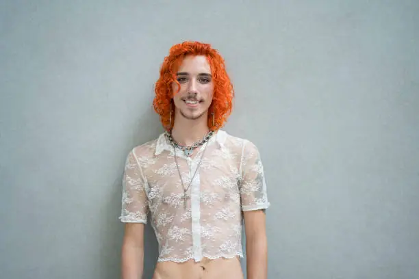 Confident individual with medium-length dyed red hair, mustache, and goatee wearing sheer summer crop top, jewelry, and smiling at camera.