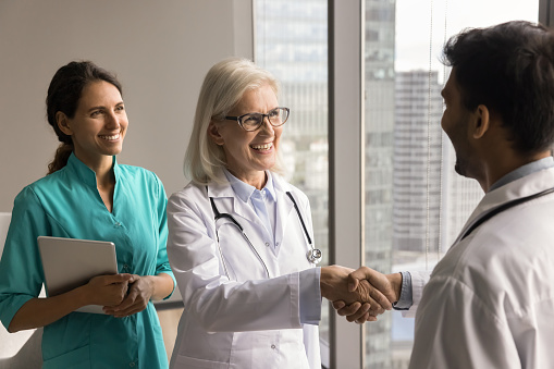 Happy senior clinic boss woman shaking hands with young male doctor, smiling, laughing, hiring new healthcare professional, expressing recognition, gratitude to younger colleague
