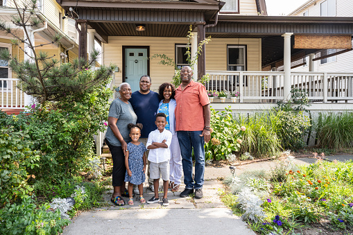 Full length view of multi-generation Black family, casually dressed and standing together in front of two-story home, smiling at camera.