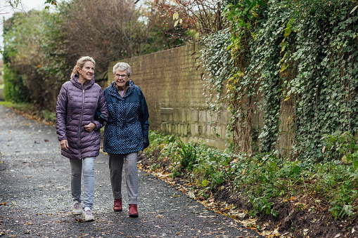 A mature woman walking with her mother on a path through a public park while getting exercise in Seghill, Northumberland. The senior woman has dementia and is arm in arm with her daughter for support and care while they talk with each other and enjoy the fresh air.\n\nVideos also available for similar scenario.