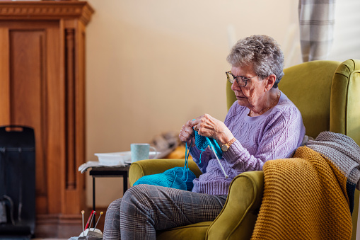 A senior woman sitting on an armchair at home in Seghill, Northumberland. She has dementia and is spending time knitting with wool and knitting needles to aid better mental health. The rhythm of knitting helps with serotonin release. This is the chemical transmitter that helps regulate anxiety, happiness, and mood. There is a strong connection between knitting and the feelings of calm and happiness in the brain. \n\nVideos also available for similar scenario.