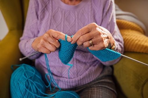 An unrecognisable senior woman sitting on an armchair in the living room at home in Seghill, Northumberland. She has dementia and is spending time knitting with wool and knitting needles to aid better mental health. The rhythm of knitting helps with serotonin release. This is the chemical transmitter that helps regulate anxiety, happiness, and mood. There is a strong connection between knitting and the feelings of calm and happiness in the brain. The main focus is her hands knitting away.

Videos also available for similar scenario.