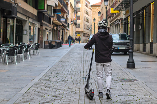 Young white guy carrying an electric scooter through the city center, with case and backpack, and people in the background.