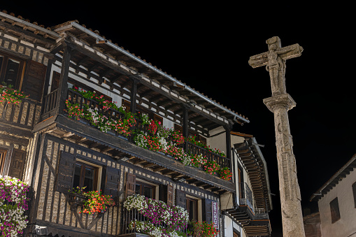 Fotografía nocturna de cruz de piedra y arquitectura tradicional con balcones de madera y tiestos con flores en la hermosa villa de La Alberca, España