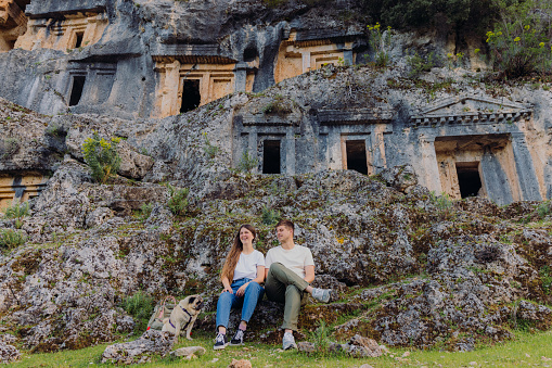 Low-angle view of a smiling woman and man with backpack sitting in the mountains contemplating views of ancient world of Pinara in South Turkey