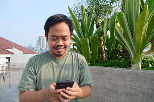 Indonesian man, wearing T-shirt,  smiling while using and looking to his smartphone in the outdoor area
