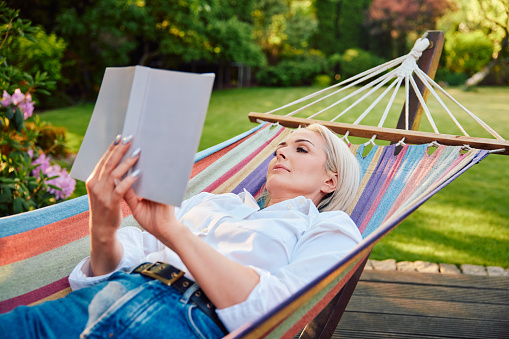 Attractive mature woman reading book in hammock relaxing in backyard