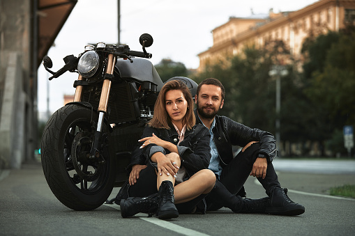 Couple of beautiful lovers are sitting hugging next to a motorcycle on the road in the city