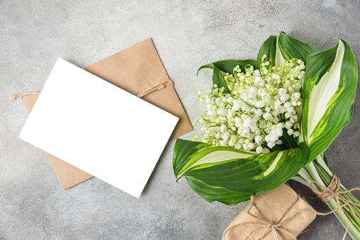 Blank greeting card with Lily of the valley flowers bouquet and gift box on gray concrete background. Wedding invitation. Womens day, Valentines day card. Mock up. Flat lay.