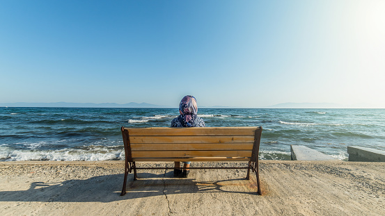 Istanbul, Turkey - 24 October 2023: An elderly muslim woman sits on a bench by the seaside, gazing at the horizon