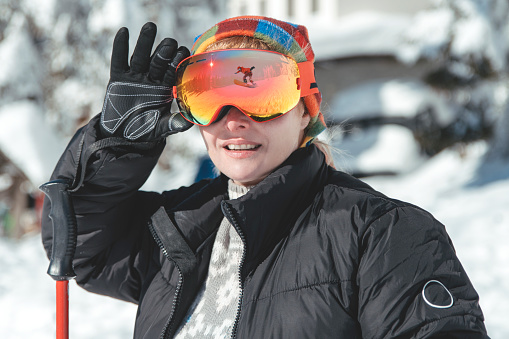 Female skier wearing ski goggles with the reflection of snowboarder