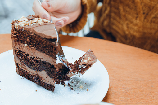 hand of unrecognizable caucasian woman cutting with fork slice of chocolate cake served on white porcelain plate on wooden table, pastry concept for social media, copy space