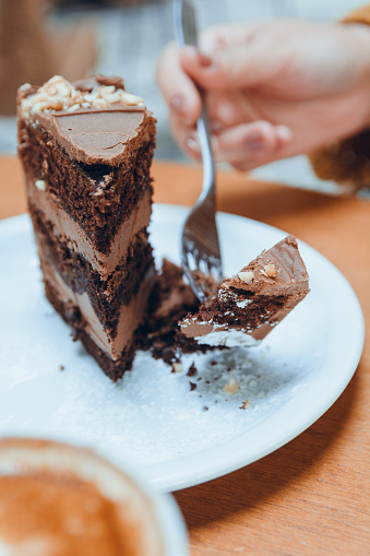 vertical image of hand of unrecognizable caucasian woman cutting with fork slice of chocolate cake served on white porcelain plate on wooden table, pastry concept for social media, copy space