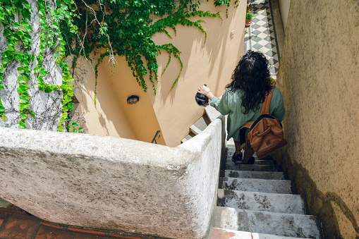 rear view of unrecognizable curly woman wearing green clothes and brown leather backpack, walking down stairs of her house, copy space.