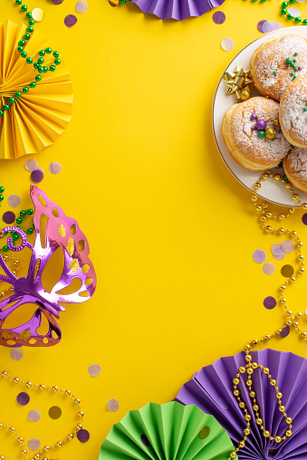 Indulgent Mardi Gras Delight: vertical top view of plate showcasing mouthwatering donuts, extravagant masquerade mask, bead necklaces, confetti on yellow backdrop. Satisfy your carnival appetite