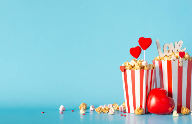 istock Valentine's Day at romantic theater. Side view reveals table adorned with striped containers loaded with caramel popcorn, heart-themed accents, love-inscribed sign, and more on light blue background 1909720874