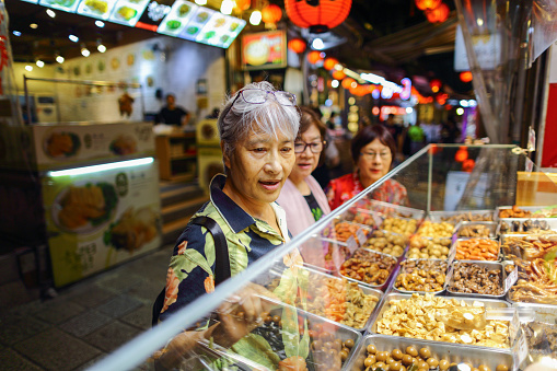 Three mature Asian women, about 70 years old, are visiting the night market in Jiufen, Taipei, which is a popular tourist attraction. They are choosing braised food and queuing up to buy it.