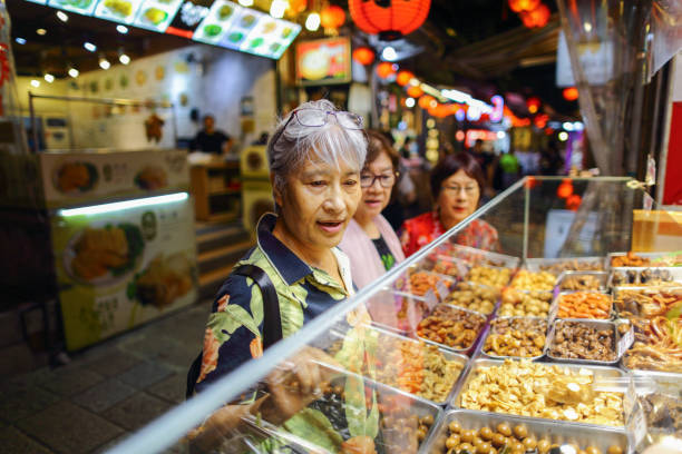 three mature asian women, about 70 years old, are visiting the night market in jiufen, taipei, which is a popular tourist attraction. they are choosing braised food and queuing up to buy it. - independence lifestyles smiling years imagens e fotografias de stock