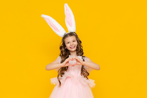 Happy Easter. A little girl dressed as a rabbit celebrates a spring holiday. A charming, smiling child in a beautiful dress with long ears on a yellow isolated background.