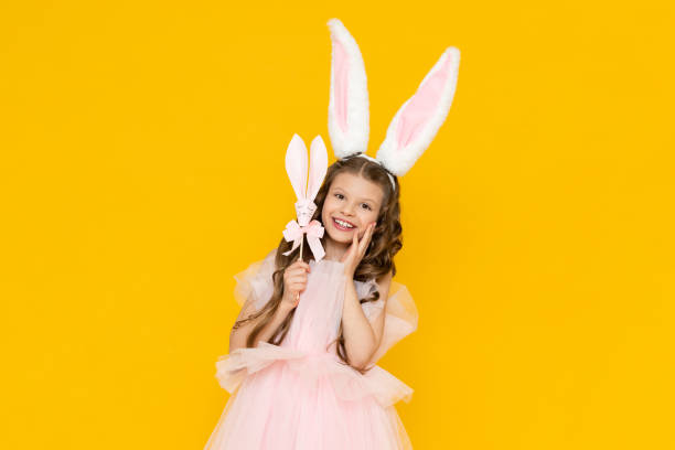 istock Happy Easter. A little girl dressed as a rabbit celebrates a spring holiday. A charming, smiling child in a beautiful dress with long ears on a yellow isolated background. 1909777408